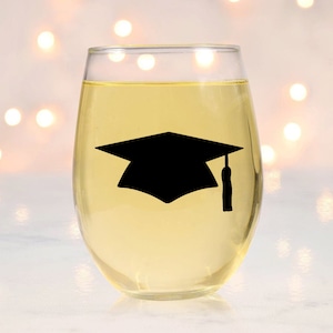 May include: A clear glass wine glass with a black graduation cap silhouette on the side. The glass is filled with a light yellow liquid.