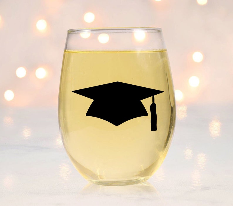 May include: A clear glass wine glass with a black graduation cap silhouette on the side. The glass is filled with a light yellow liquid.