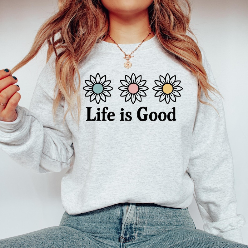 May include: A light grey sweatshirt featuring the phrase "Life is Good" in black lettering. Above the text are three daisy flowers with pastel blue, pink, and yellow centres. The sweatshirt is a casual, comfortable style.