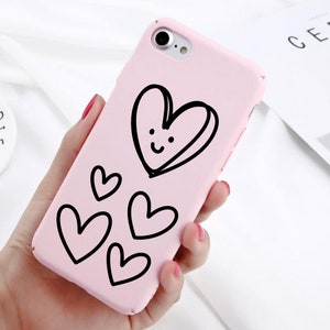 May include: A pink phone case with a black outline of a heart with a smiley face and smaller hearts around it.