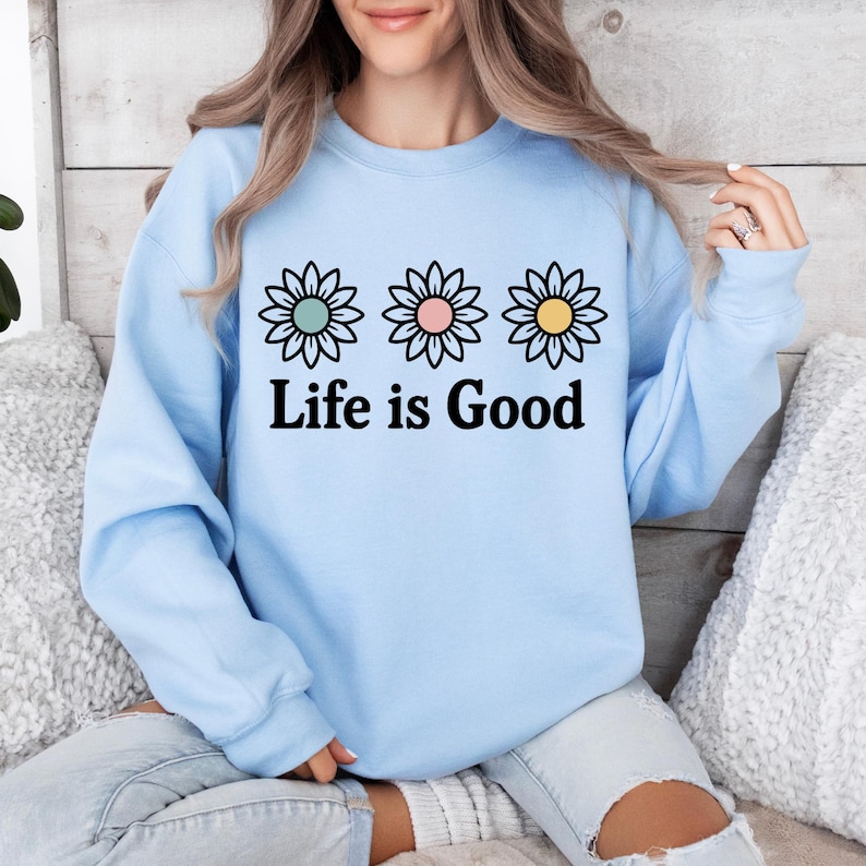 May include: A pale blue sweatshirt displaying three daisy designs with pastel centres and black outlines. The text "Life is Good" is printed in black beneath the flowers. The sweatshirt is a casual, comfortable style.