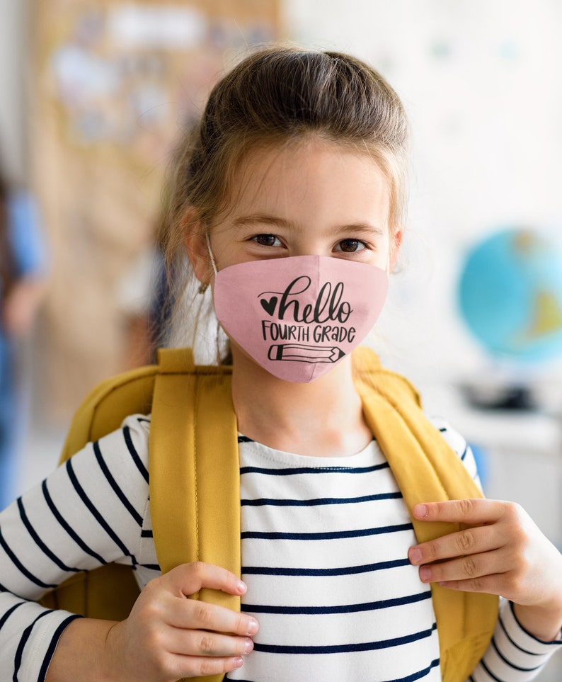 以下が含まれることがあります： ピンクのマスクをつけた若い人が、「hello fourth grade」という文字とハートと鉛筆のデザインをしています。ストライプのシャツと黄色のバックパックを着用しています。