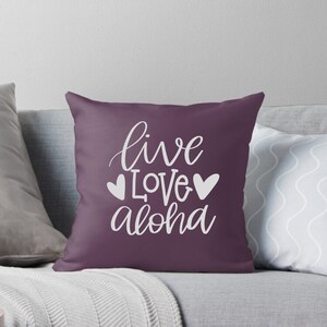 May include: Purple throw pillow with white text that reads "live love aloha" with two white hearts on either side of the word "love".