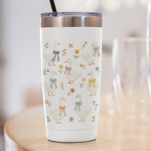 May include: White travel tumbler with a silver lid and a black straw. The tumbler is decorated with illustrations of white geese wearing colorful bows and surrounded by flowers. The design is repeated around the tumbler.