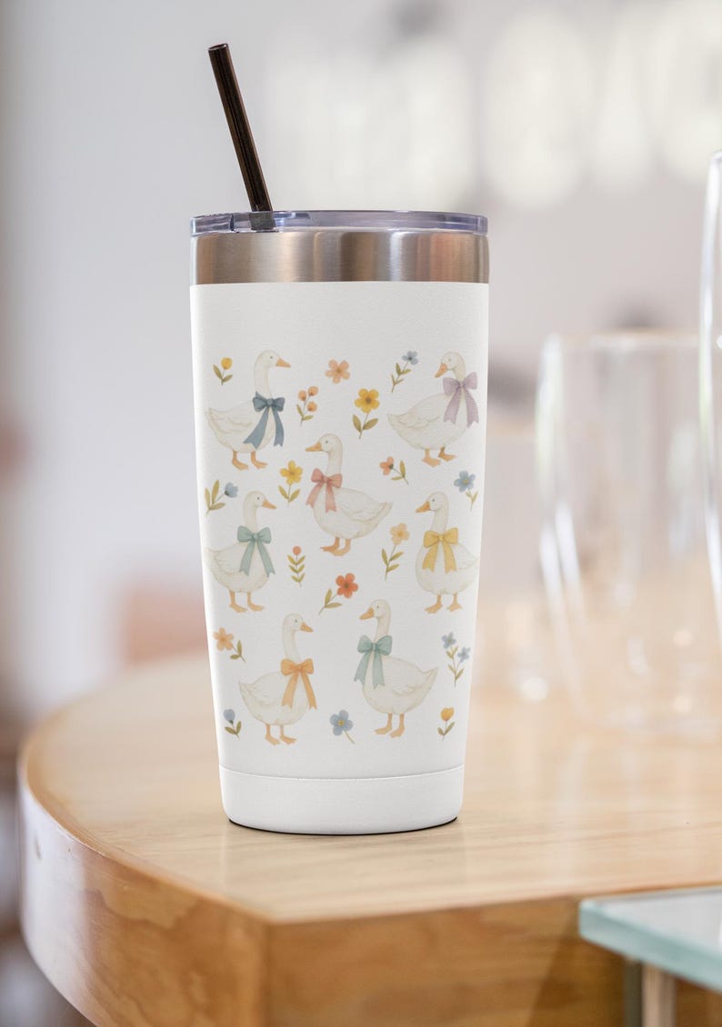 May include: White travel tumbler with a silver lid and a black straw. The tumbler is decorated with illustrations of white geese wearing colorful bows and surrounded by flowers. The design is repeated around the tumbler.