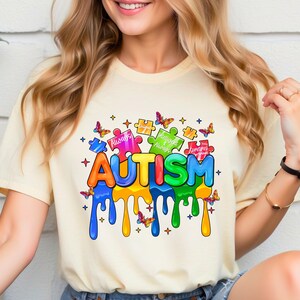 Pode incluir: Uma camiseta amarela com um gr&aacute;fico colorido da palavra "AUTISM" com pe&ccedil;as de quebra-cabe&ccedil;a e borboletas. As pe&ccedil;as do quebra-cabe&ccedil;a t&ecirc;m as palavras "&Uacute;nico", "Sempre", "Inteligente" e "Torna-se" nelas.