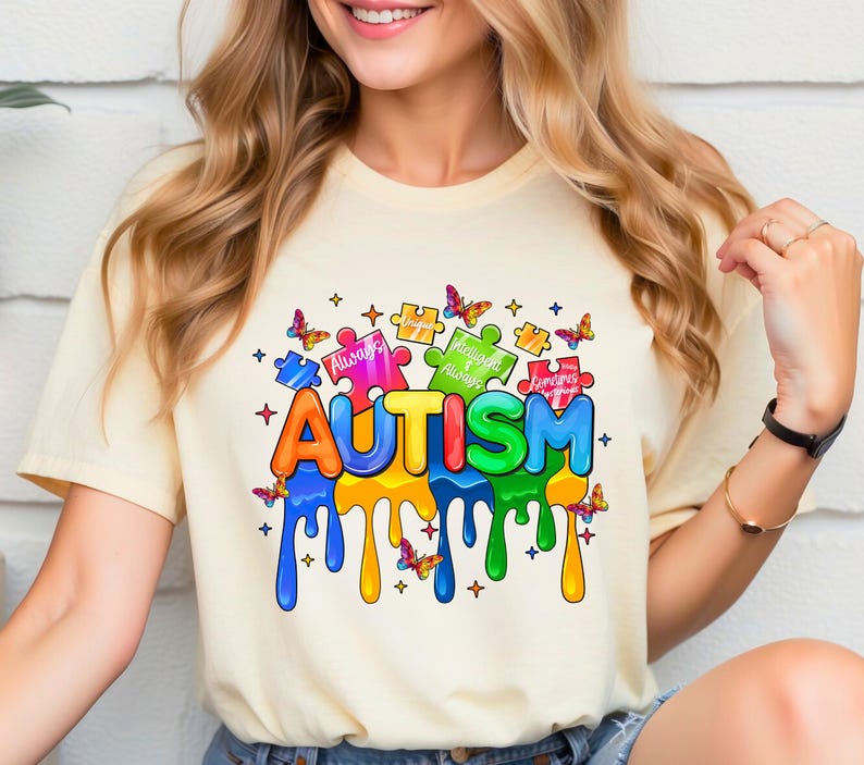 Pode incluir: Uma camiseta amarela com um gr&aacute;fico colorido da palavra "AUTISM" com pe&ccedil;as de quebra-cabe&ccedil;a e borboletas. As pe&ccedil;as do quebra-cabe&ccedil;a t&ecirc;m as palavras "&Uacute;nico", "Sempre", "Inteligente" e "Torna-se" nelas.