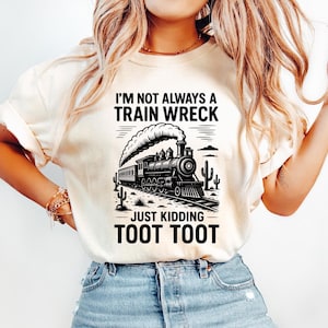 May include: Cream-colored t-shirt featuring a black and white illustration of a vintage train with smoke billowing from its smokestack. The text reads "I'M NOT ALWAYS A TRAIN WRECK JUST KIDDING TOOT TOOT."