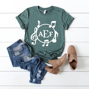 May include: A green t-shirt with a white circle design featuring musical notes and the letters "AEF".