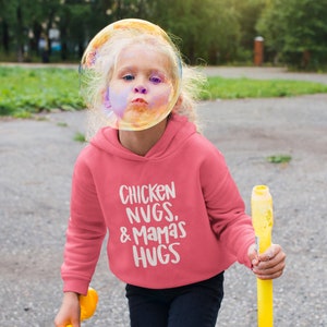 May include: A pink hoodie with the text "Chicken Nugs. & Mamas Hugs" in white. A young person is wearing the hoodie and holding a yellow bubble wand.