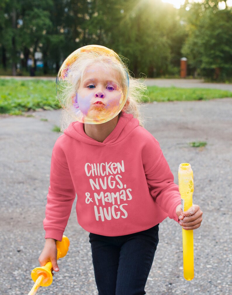 May include: A pink hoodie with the text "Chicken Nugs. & Mamas Hugs" in white. A young person is wearing the hoodie and holding a yellow bubble wand.