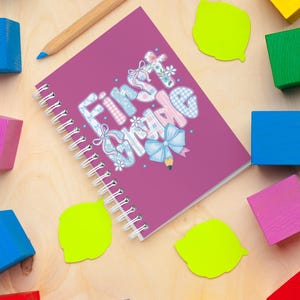 May include: A spiral-bound notebook with the words "First Grade" in a whimsical font, decorated with bows and flowers. A wooden pencil and colorful blocks surround the notebook on a light wood surface. Bright green sticky notes add a pop of color.