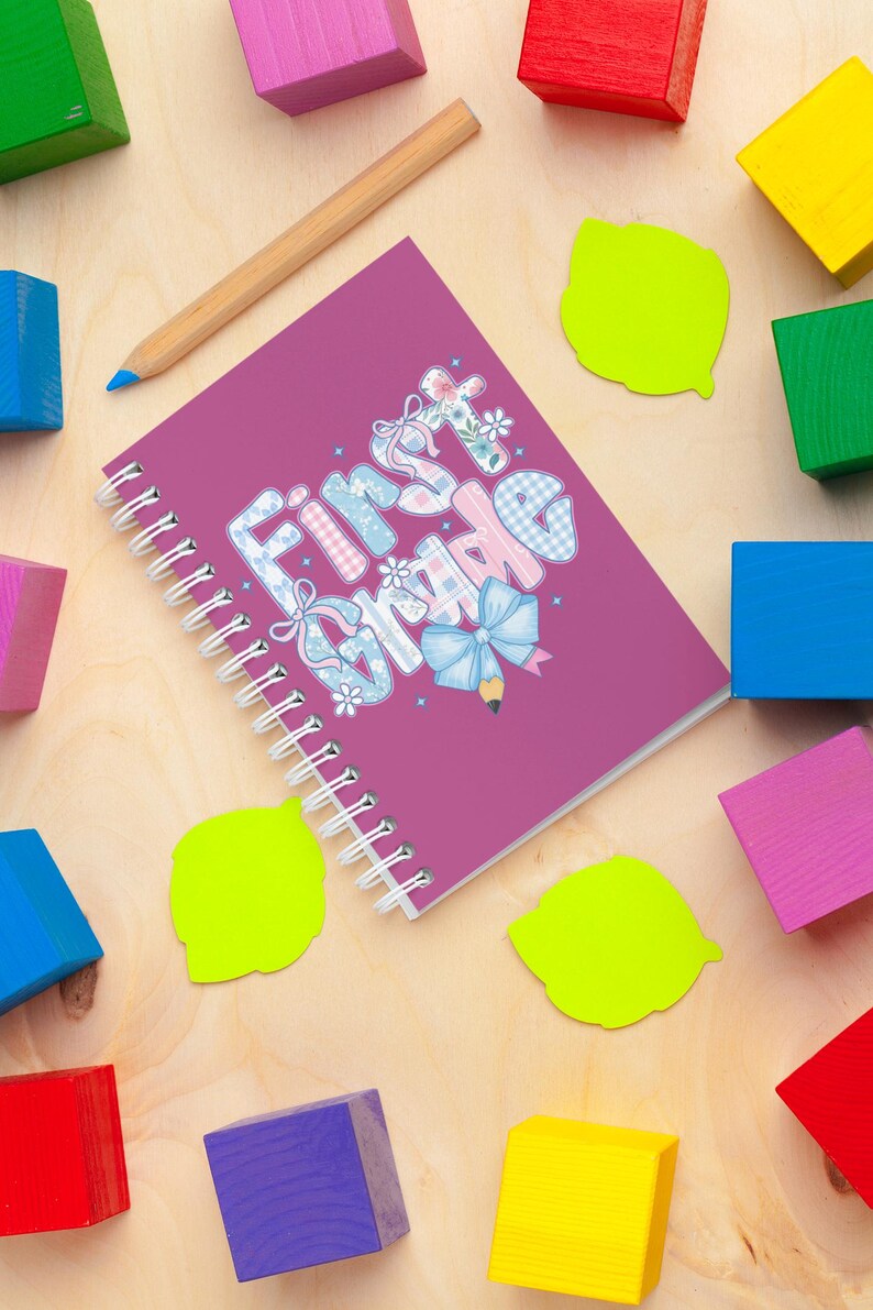 May include: A spiral-bound notebook with the words "First Grade" in a whimsical font, decorated with bows and flowers. A wooden pencil and colorful blocks surround the notebook on a light wood surface. Bright green sticky notes add a pop of color.