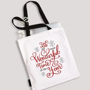 May include: White canvas tote bag with black handles. The bag has red text that says "It's the most Wonderful Time of the Year" with grey snowflakes.