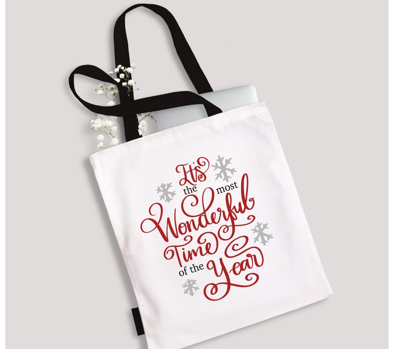 May include: White canvas tote bag with black handles. The bag has red text that says "It's the most Wonderful Time of the Year" with grey snowflakes.