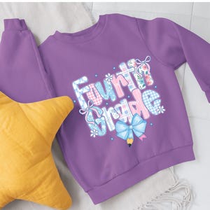 May include: A purple sweatshirt with the words "Fourth Grade" in a colorful, whimsical design. The text incorporates floral patterns, bows, and a pencil graphic. A yellow star-shaped pillow is in the foreground.