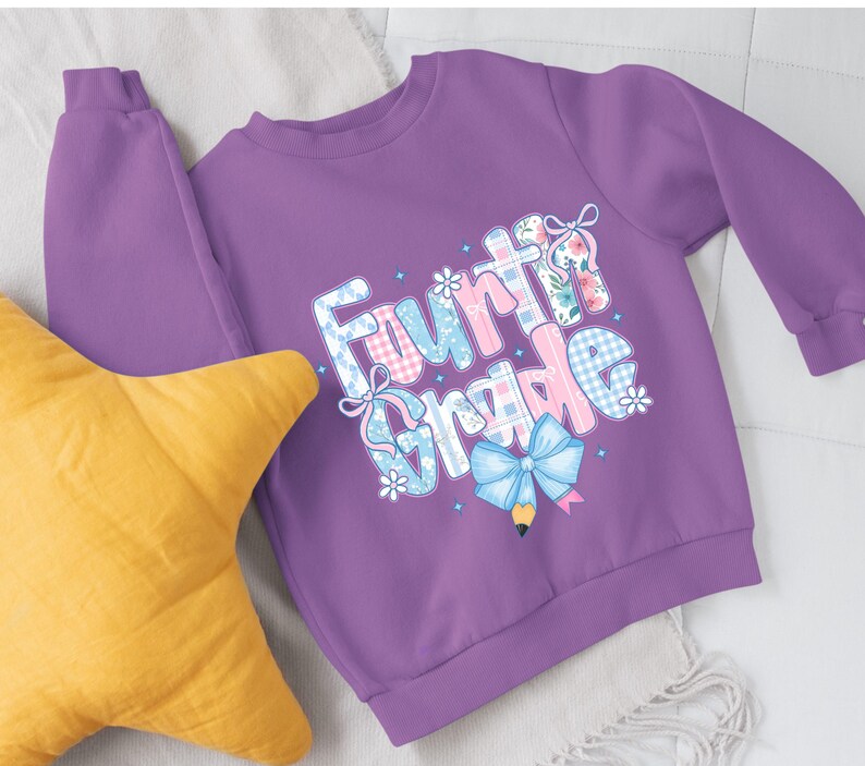 May include: A purple sweatshirt with the words "Fourth Grade" in a colorful, whimsical design. The text incorporates floral patterns, bows, and a pencil graphic. A yellow star-shaped pillow is in the foreground.
