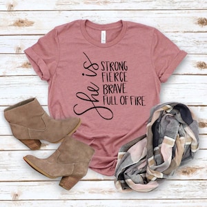 May include: A mauve t-shirt with the text "She is strong, fierce, brave, full of fire" printed on the front. The shirt is paired with a brown suede ankle boot and a plaid scarf.