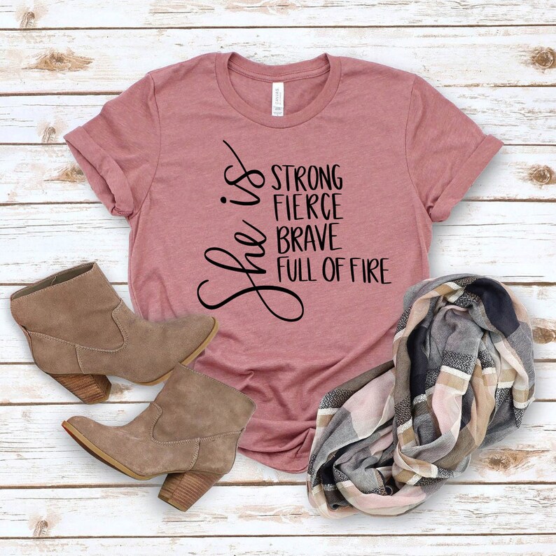 May include: A mauve t-shirt with the text "She is strong, fierce, brave, full of fire" printed on the front. The shirt is paired with a brown suede ankle boot and a plaid scarf.