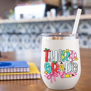 Puede incluir: Un vaso de vino de acero inoxidable blanco con un gr&aacute;fico colorido que dice "Tercer Grado". El gr&aacute;fico incluye una manzana roja, un globo terr&aacute;queo azul, un lazo rosa y l&aacute;pices amarillos.