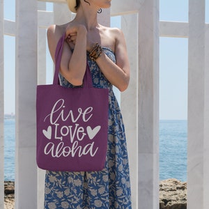 May include: A woman is holding a purple tote bag with the text "live love aloha" in white lettering and two white hearts on a beach.