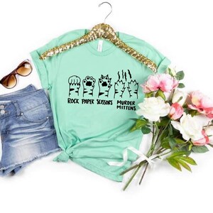 May include: A light green t-shirt with a graphic of cat paws in the shape of rock, paper, scissors, and murder mittens. The text "Rock Paper Scissors Murder Mittens" is printed below the graphic.