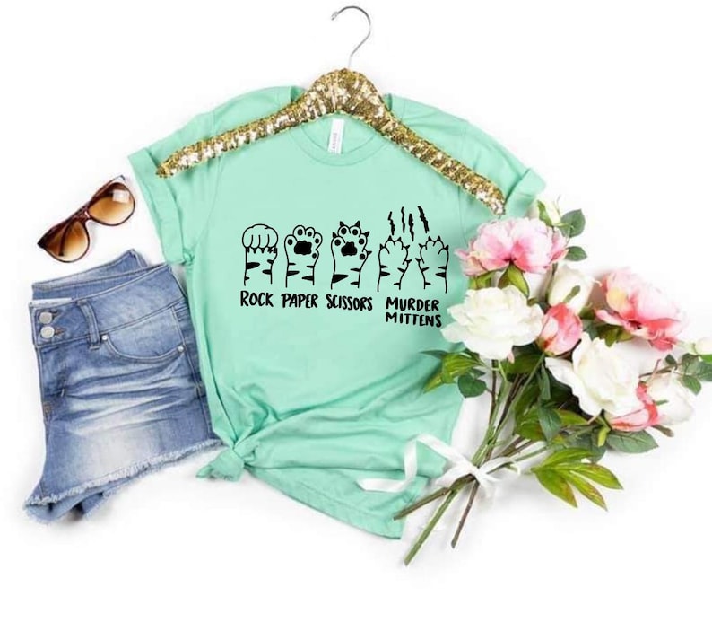 May include: A light green t-shirt with a graphic of cat paws in the shape of rock, paper, scissors, and murder mittens. The text "Rock Paper Scissors Murder Mittens" is printed below the graphic.