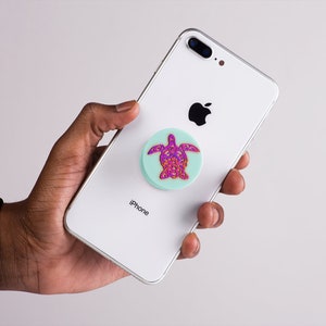 K&ouml;nnte beinhalten: Ein wei&szlig;es iPhone mit einem t&uuml;rkisfarbenen und rosafarbenen Schildkr&ouml;ten-Handyhalter, der an der R&uuml;ckseite befestigt ist. Das Telefon tr&auml;gt auf der R&uuml;ckseite die Aufschrift "iPhone".
