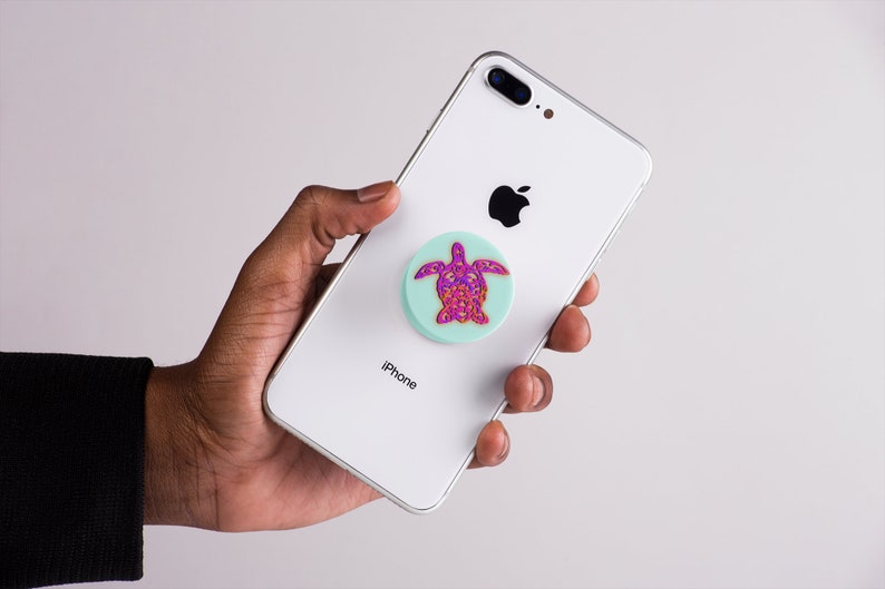 K&ouml;nnte beinhalten: Ein wei&szlig;es iPhone mit einem t&uuml;rkisfarbenen und rosafarbenen Schildkr&ouml;ten-Handyhalter, der an der R&uuml;ckseite befestigt ist. Das Telefon tr&auml;gt auf der R&uuml;ckseite die Aufschrift "iPhone".