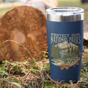 May include: Navy blue insulated tumbler with a silver rim, featuring the text "Protect Our National Parks" and a graphic of a mountain scene and a fish. The tumbler is cylindrical and appears to be made of stainless steel.