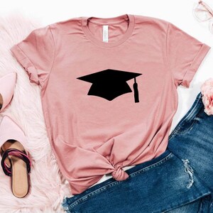 May include: A pink t-shirt with a black graduation cap graphic. The shirt is laid out on a white surface with a pair of pink shoes and blue jeans.