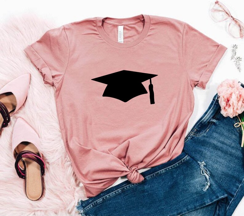 May include: A pink t-shirt with a black graduation cap graphic. The shirt is laid out on a white surface with a pair of pink shoes and blue jeans.