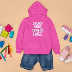May include: Pink hooded sweatshirt with the text "Chicken Nugs, & Mamas Hugs" in white. The sweatshirt is paired with blue denim shorts and pink sneakers.