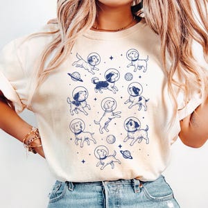 May include: A cream-coloured t-shirt with a repeating pattern of cartoon dogs wearing space helmets, planets, and stars in blue. The dogs are in various poses, creating a playful design. The shirt has short sleeves and a relaxed fit.