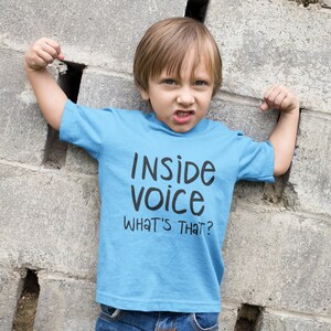 May include: A young boy wearing a light blue t-shirt with the text "Inside voice what's that?" printed on it. He is standing in front of a grey cinder block wall.