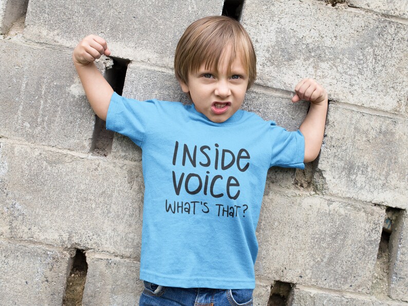 May include: A young boy wearing a light blue t-shirt with the text "Inside voice what's that?" printed on it. He is standing in front of a grey cinder block wall.