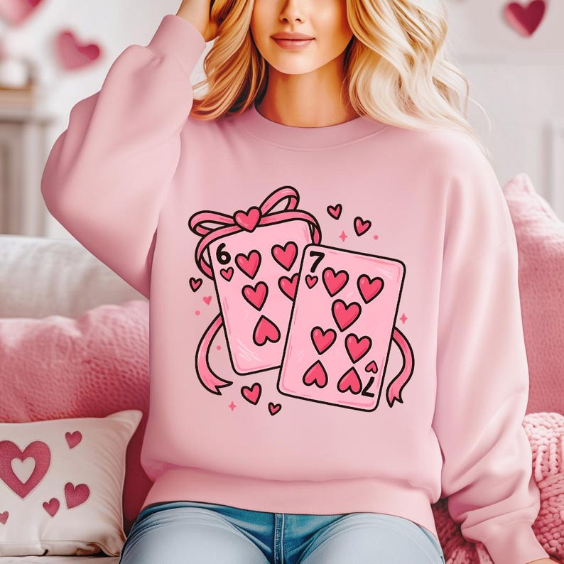 May include: A light pink sweatshirt featuring a graphic of two playing cards with hearts. The cards are tied with a pink ribbon and have the numbers 6 and 7. Small pink hearts and stars are scattered around the cards.
