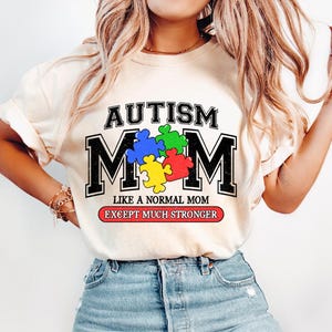 May include: A white t-shirt with the text "AUTISM MOM" in black letters with a puzzle piece design. The text "LIKE A NORMAL MOM EXCEPT MUCH STRONGER" is printed in red and black below the main text.