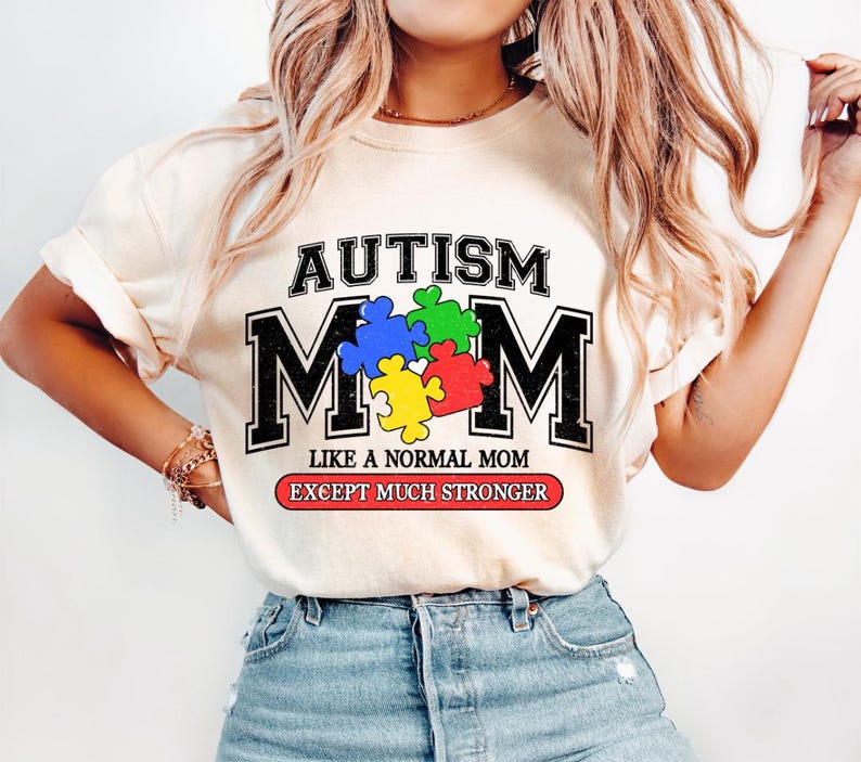 May include: A white t-shirt with the text "AUTISM MOM" in black letters with a puzzle piece design. The text "LIKE A NORMAL MOM EXCEPT MUCH STRONGER" is printed in red and black below the main text.