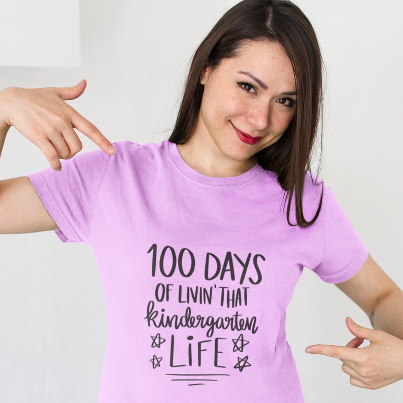 May include: A woman wearing a light purple t-shirt with the text "100 Days of Livin' That Kindergarten Life" printed on it. The text is in black and has a handwritten style.