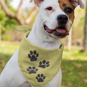 May include: A white and brown dog wearing a yellow bandana with black paw prints. The dog is sitting in a grassy area with trees in the background.