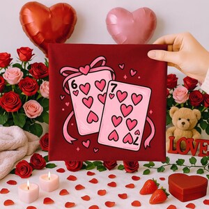 May include: A red square canvas featuring two playing cards with heart symbols, a six and a seven of hearts. The canvas is held by a hand, surrounded by roses, candles, strawberries, and heart-shaped balloons, creating a romantic theme.