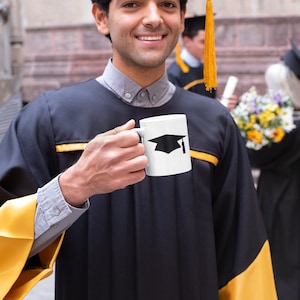 May include: A person wearing a black graduation gown with gold trim holds a white coffee mug with a black graduation cap design. The person is smiling and looking at the camera.