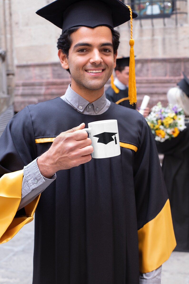 May include: A person wearing a black graduation gown with gold trim holds a white coffee mug with a black graduation cap design. The person is smiling and looking at the camera.