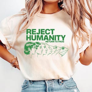 May include: Cream-colored t-shirt with the green text "REJECT HUMANITY RETURN TO FROG." The graphic features a line of human profiles morphing into a frog. The shirt is made of a soft material and has short sleeves.