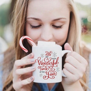 May include: White ceramic mug with a red and grey design that says "It's the most Wonderful Time of the Year". The mug is filled with a hot drink and topped with a marshmallow. A red and white candy cane is in the background.