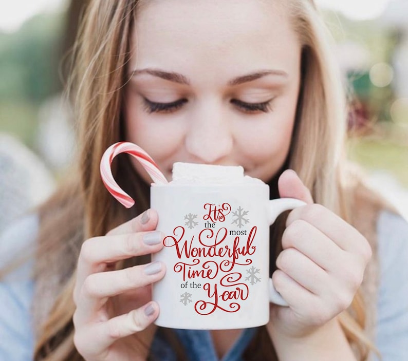 May include: White ceramic mug with a red and grey design that says "It's the most Wonderful Time of the Year". The mug is filled with a hot drink and topped with a marshmallow. A red and white candy cane is in the background.