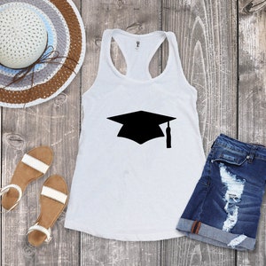 May include: White tank top with a black graduation cap graphic. The tank top is laying on a wooden surface with a straw hat, sandals, and denim shorts.