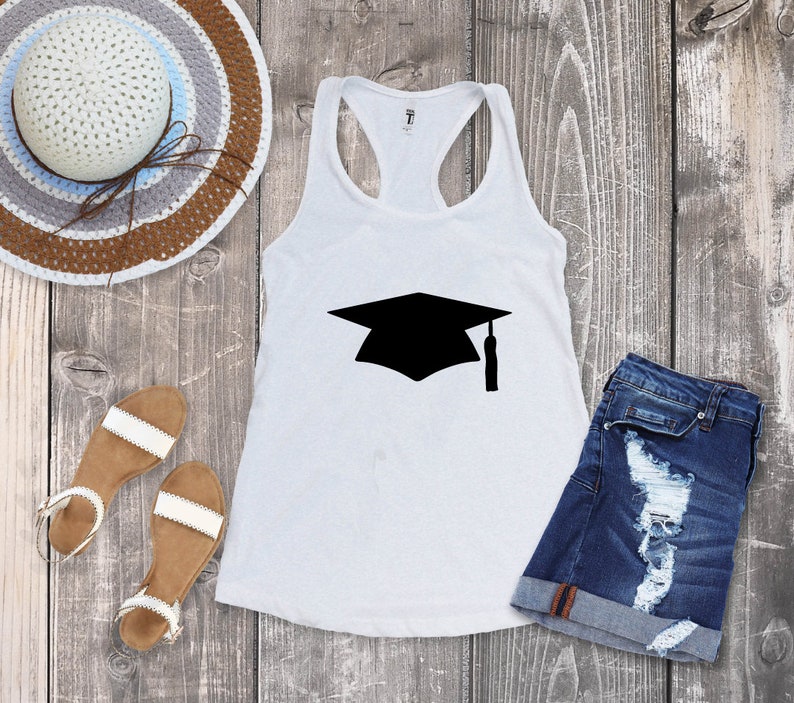 May include: White tank top with a black graduation cap graphic. The tank top is laying on a wooden surface with a straw hat, sandals, and denim shorts.