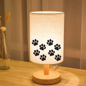 May include: A white linen lampshade with black paw prints, a wooden base, and a wooden stem. The lamp is on a wooden table.